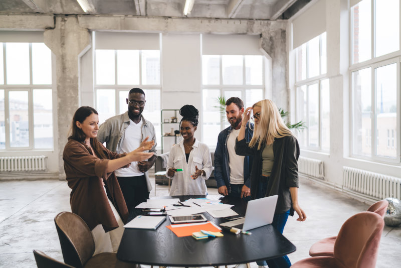 Modern young diverse women and men standing at table in spacious light loft office and talking with inspiration while coworking on new business startup